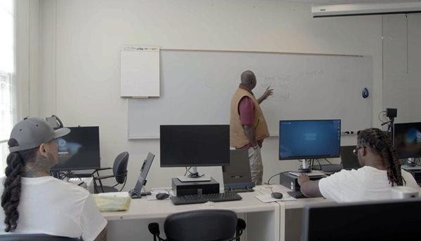 Students at computers listen to an instructor at a whiteboard